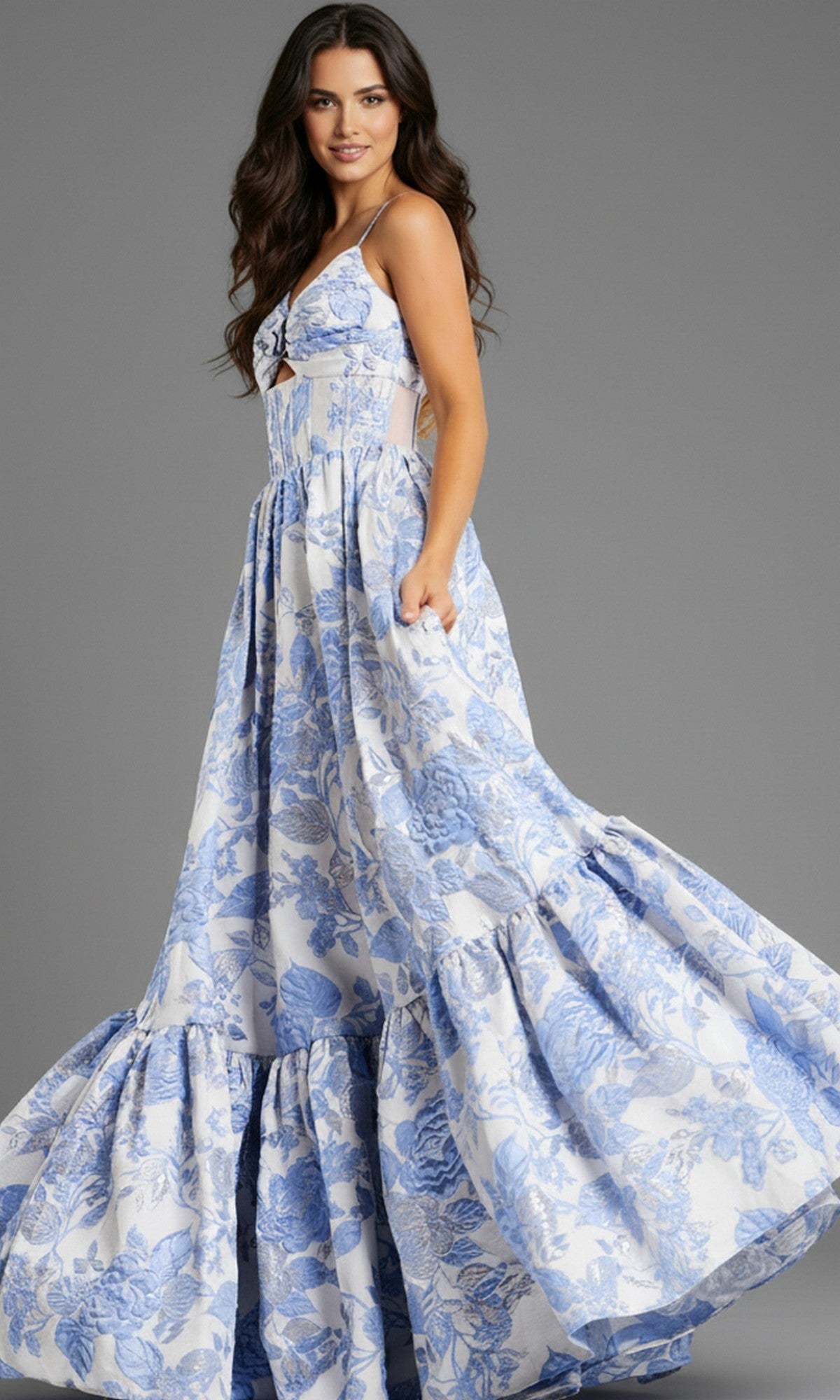A woman stands against a plain gray background wearing the Jovani 47328, a long, sleeveless blue and white floral jacquard prom dress with an A-line skirt.